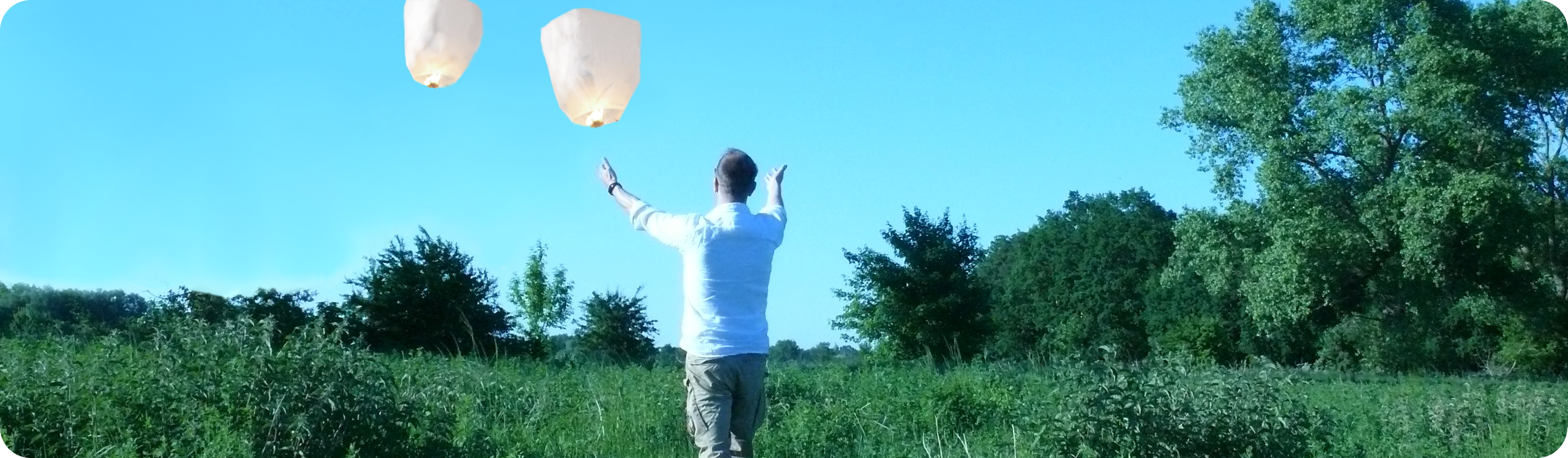 Grüne landschaft mit hellblauem himmel eine Person mit geöffneten Armen zum Himmel und zwei davon fliegende Himmellaternen